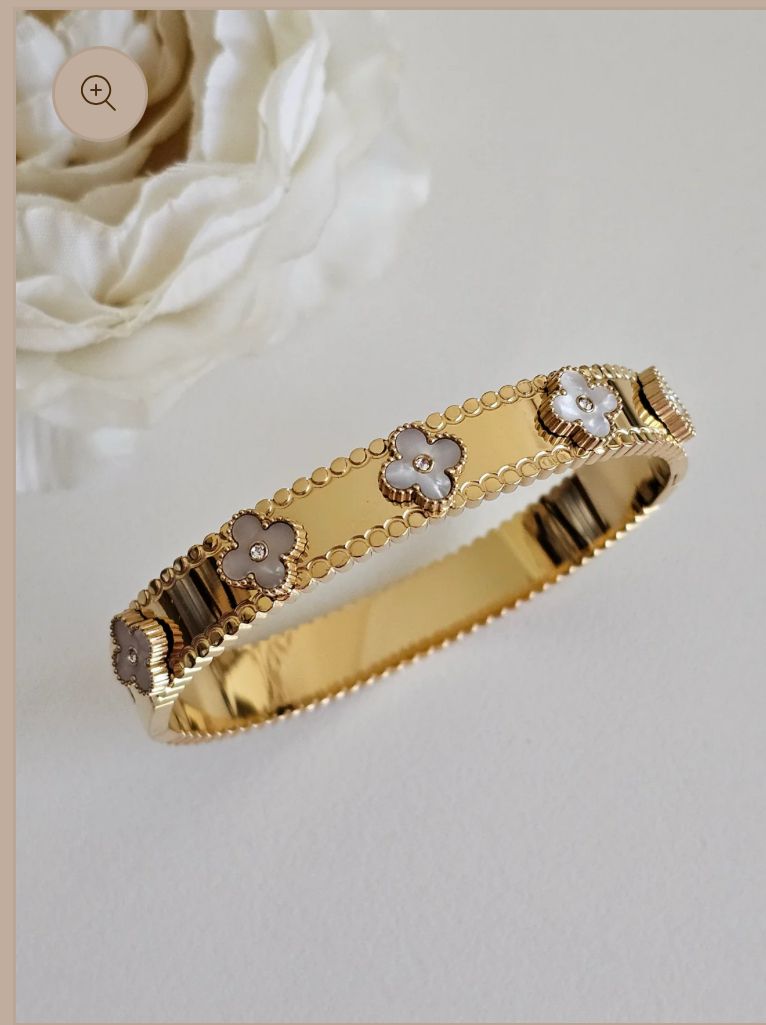An 18k gold plated helen clover white bangle with a clover pattern adorned with zirconia, displayed against a floral background.