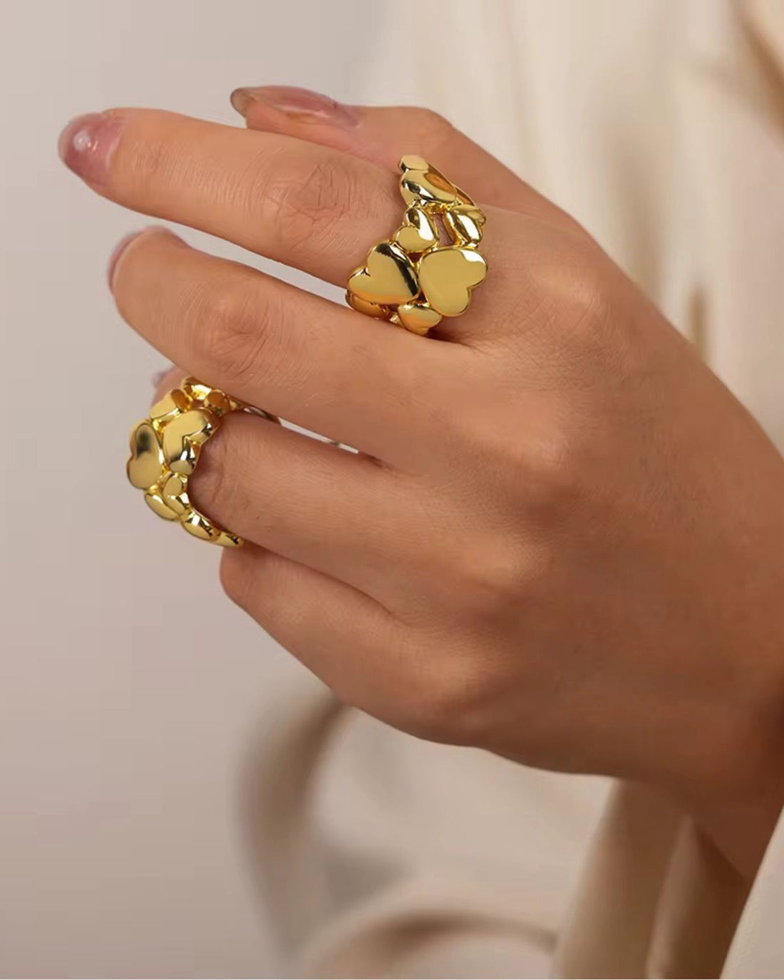Hand wearing two gold gloria heart rings with a plain background