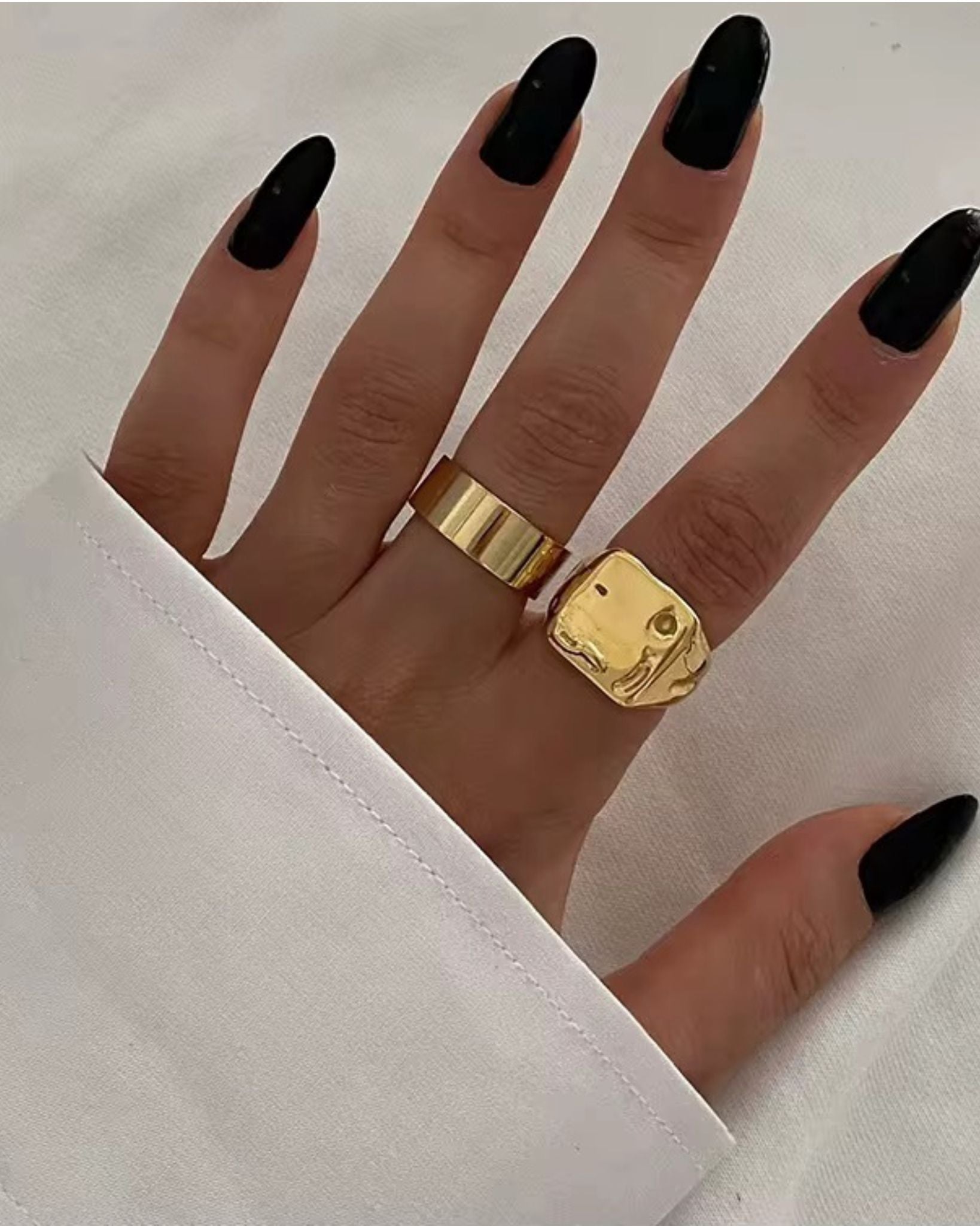 Hand wearing two gold goldy rings with black nail polish on a white background