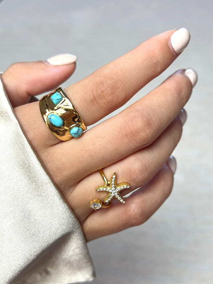 Hand wearing two gold rings with turquoise stones and a starfish design on a neutral background