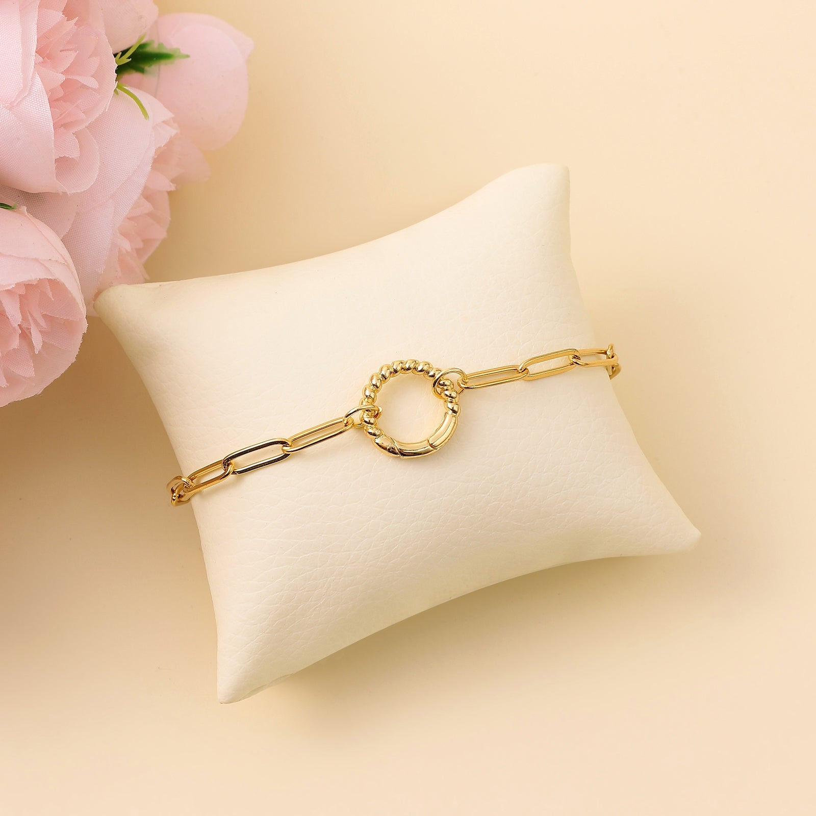 An 18k gold plated bracelet with a round clasp, displayed on a white cushion, with pink flowers and a hand partially visible in the background.